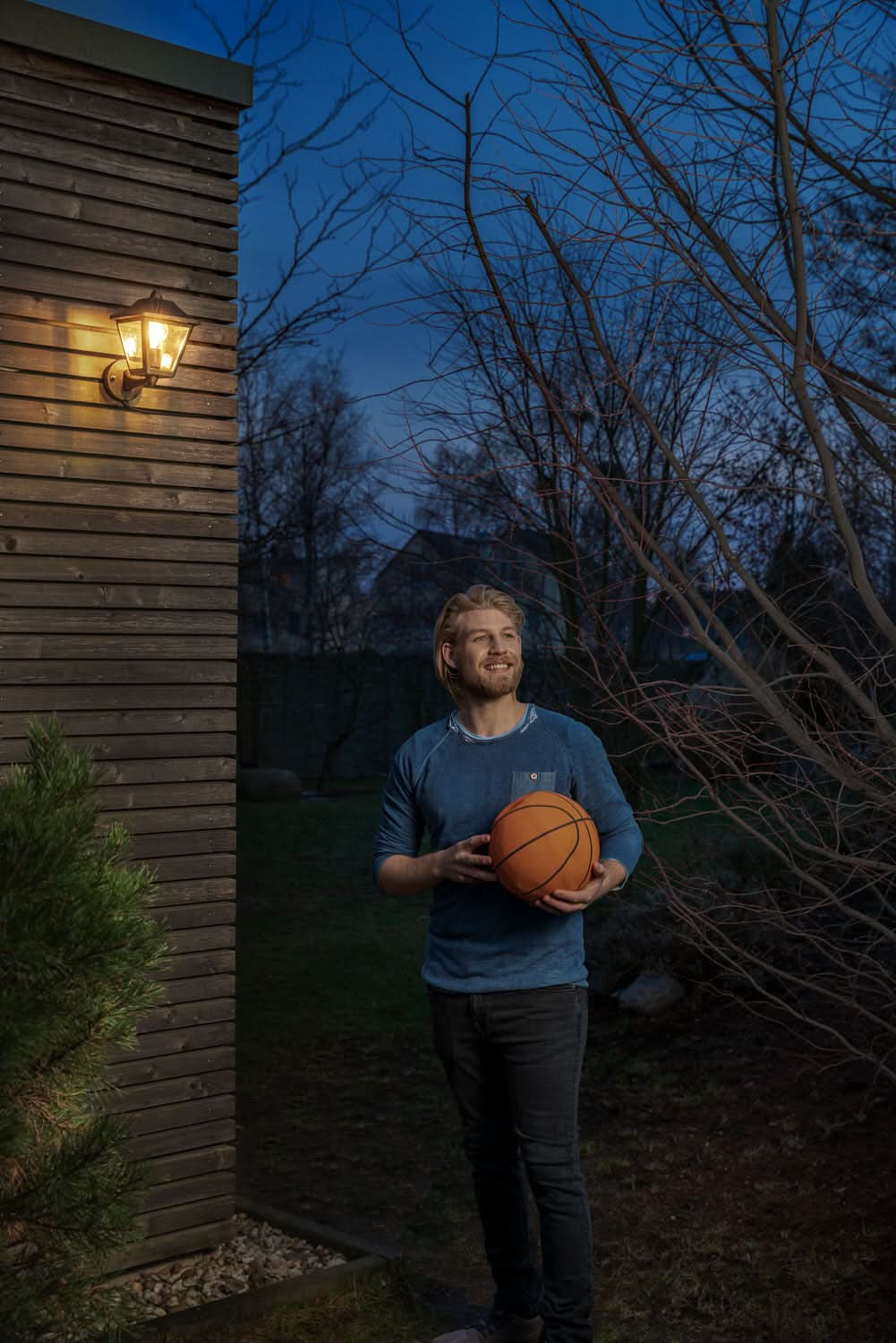 Homme au ballon devant un mur boisé, applique allumée dans un jardin au crépuscule.