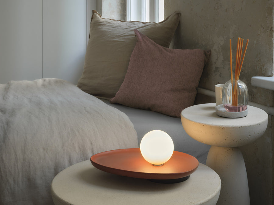 Lampe allumée sur table d'appoint, près d'un lit et de coussins beiges.