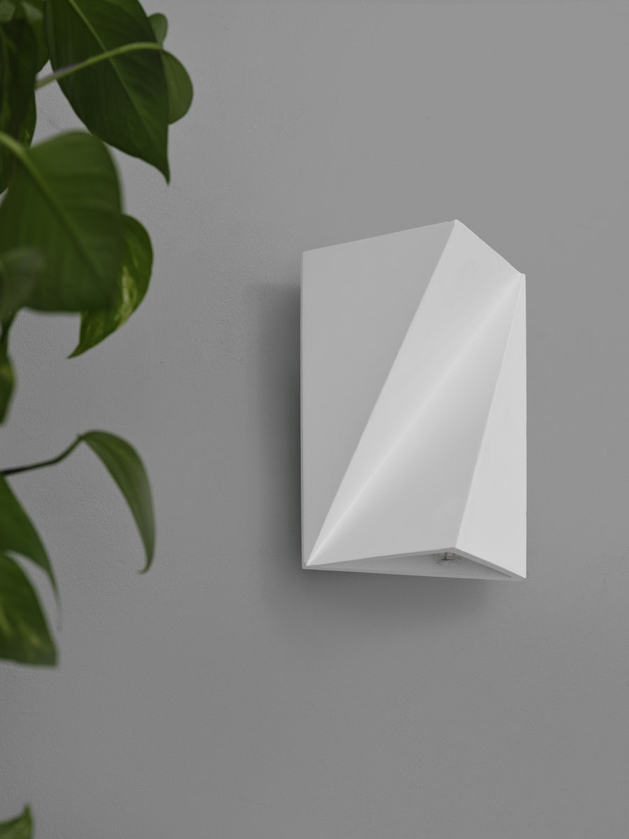 Face avant de l'applique blanche montée sur mur gris, avec feuillage flou au premier plan.