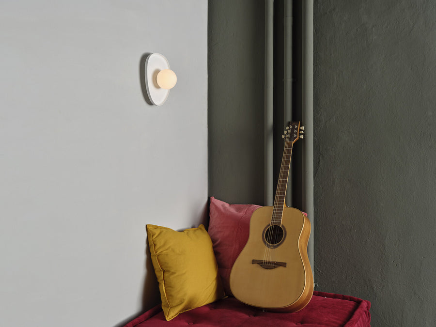Applique murale allumée sur un mur gris, près d’une guitare et de coussins colorés.