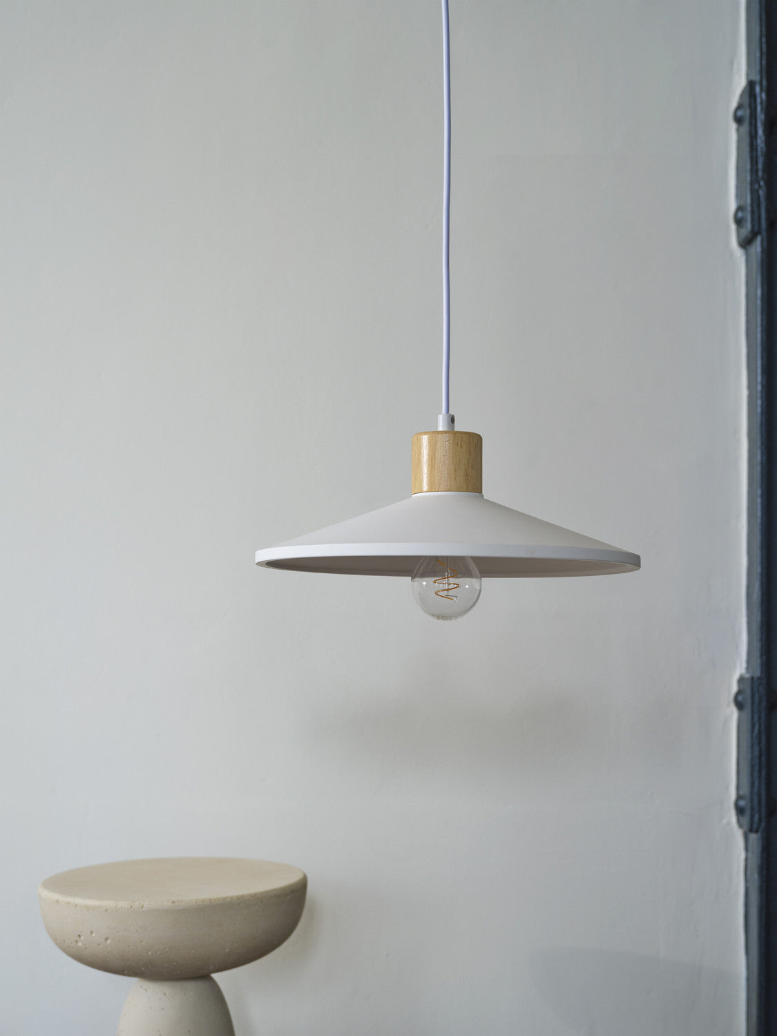 Suspension blanche éclairée, ampoule apparente, devant un mur neutre et un tabouret béton.