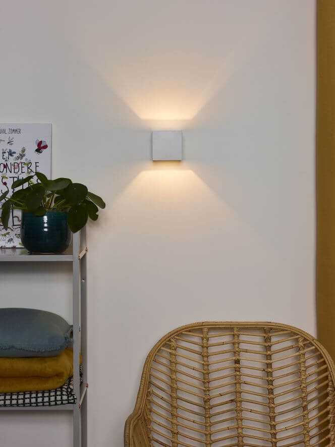 Vue rapprochée de l’applique blanche allumée, avec une plante verte et une étagère à gauche.