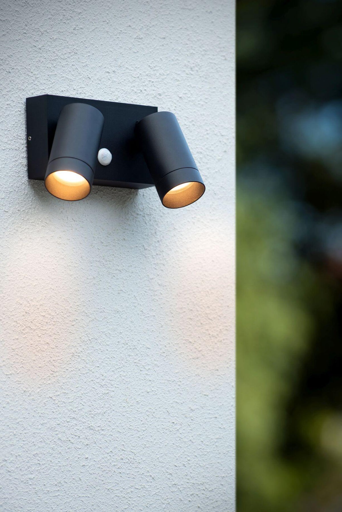 Applique murale noire allumée sur mur crépis, deux faisceaux lumineux et détecteur.