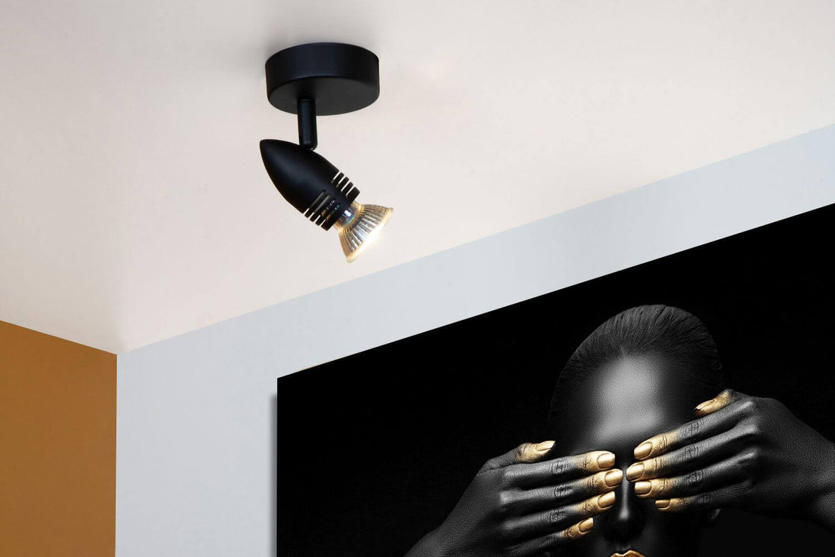 Vue rapprochée du spot plafonnier noir allumé devant un tableau noir et or.