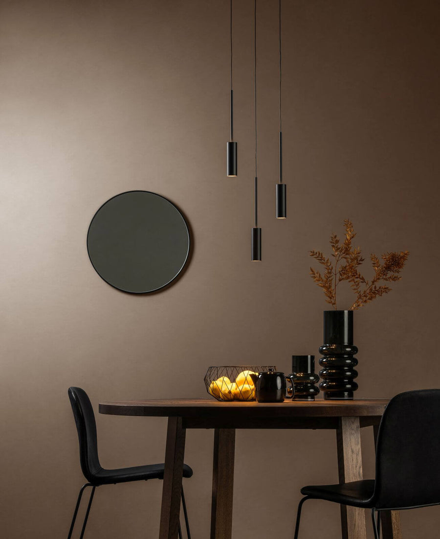 Suspension noire à trois tubes éclairant une table en bois dans un intérieur chaleureux.