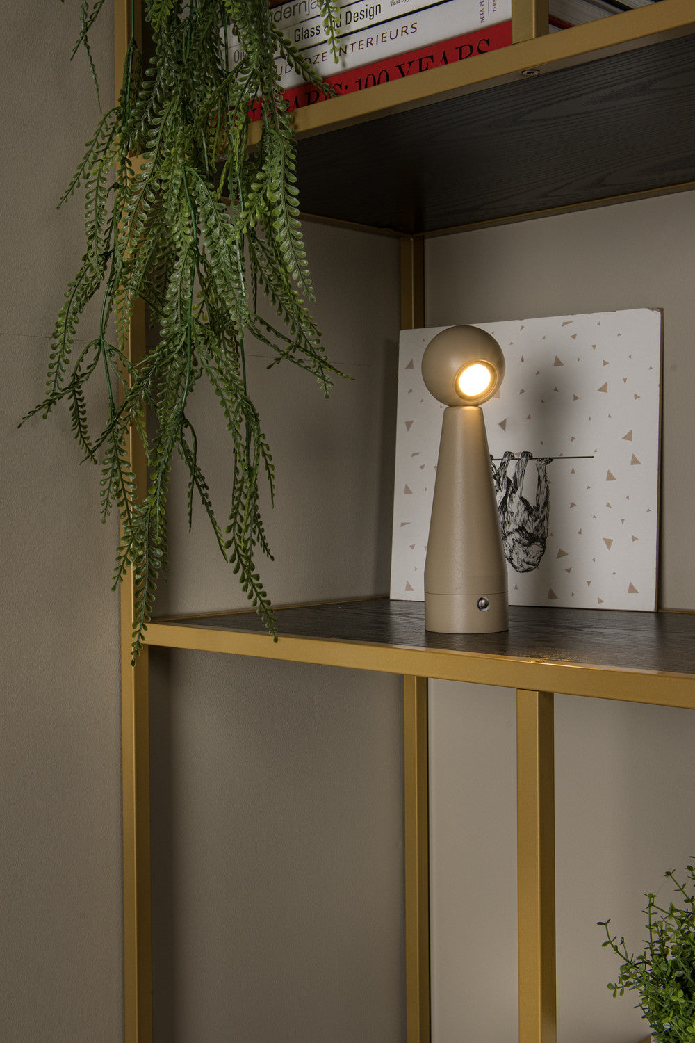 Mise en ambiance du luminaire posé parmi livres et verdure sur étagère dorée.