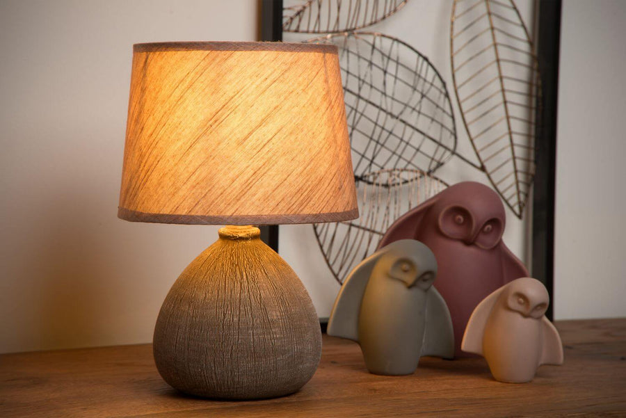 Lampe à poser allumée devant un fond décoratif, avec des chouettes en céramique sur bois.
