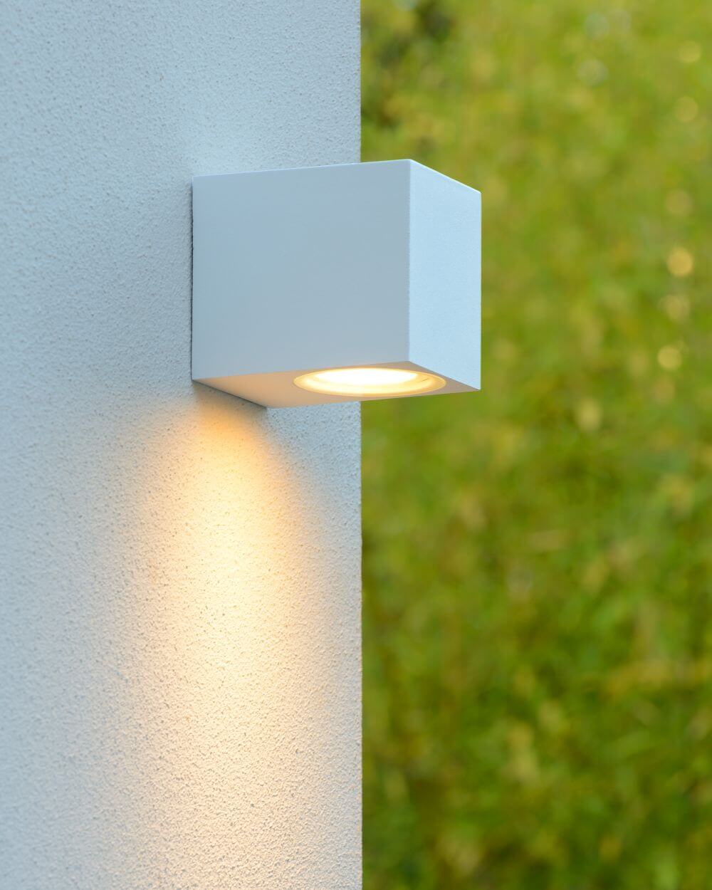 Applique murale blanche allumée sur un mur extérieur, halo lumineux vers le bas.