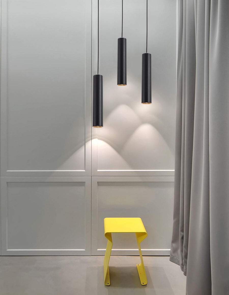 Trois suspensions cylindriques noires éclairant un mur gris, tabouret jaune en dessous
