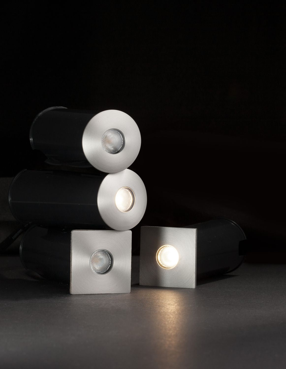Trois luminaires encastrables posés sur fond noir, montrant versions rondes et carrées éclairées.
