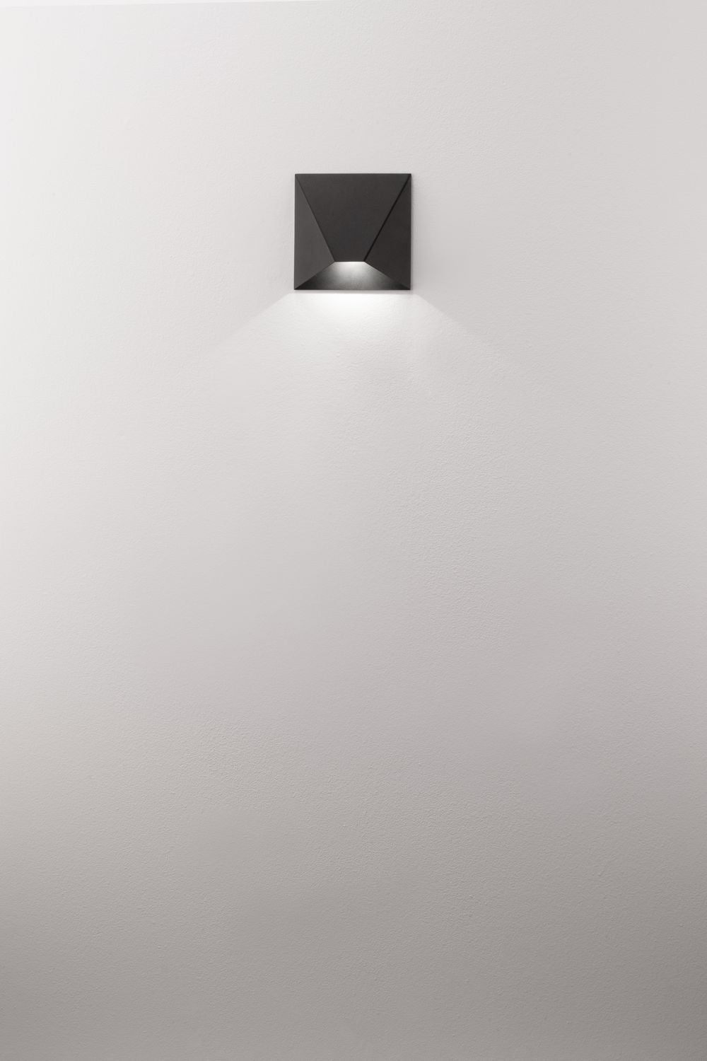 Luminaire mural noir allumé sur mur blanc, halo diffusant une lumière rasante vers le sol