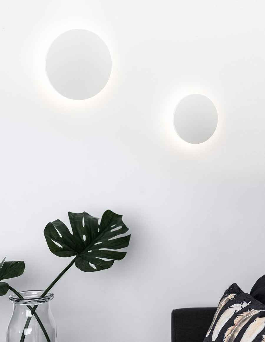 Deux appliques rondes blanches allumées sur mur blanc, plante et coussin visibles
