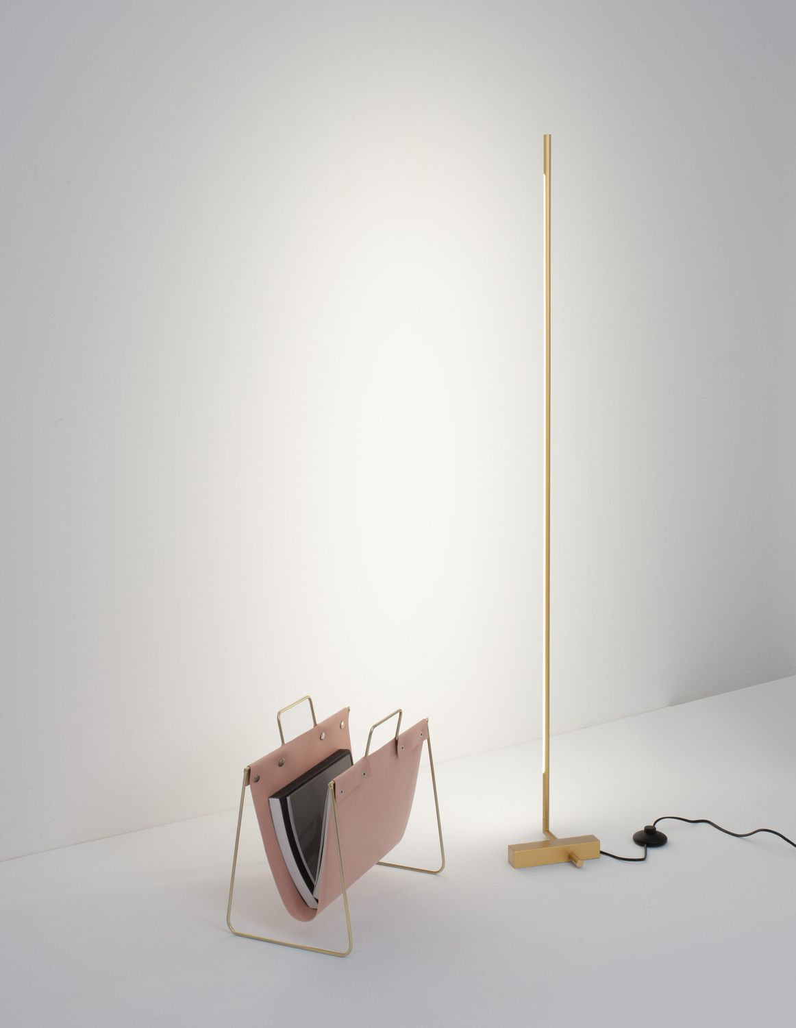 Lampadaire laiton fin et haut, à côté d'un porte-revues rose dans un intérieur blanc.
