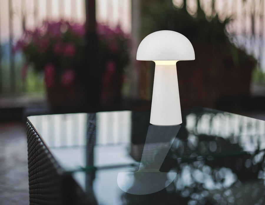 Lampe blanche posée sur table en verre, reflet visible, fleurs floues en arrière-plan