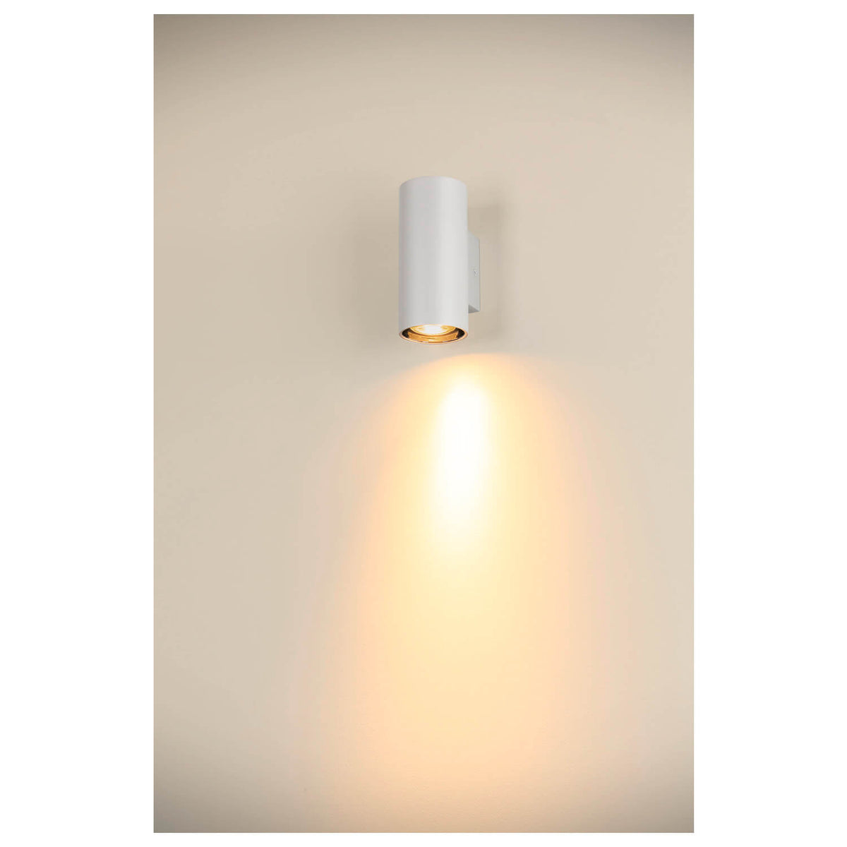 Mise en ambiance sur un mur beige, halo lumineux descendant sous l’applique blanche.