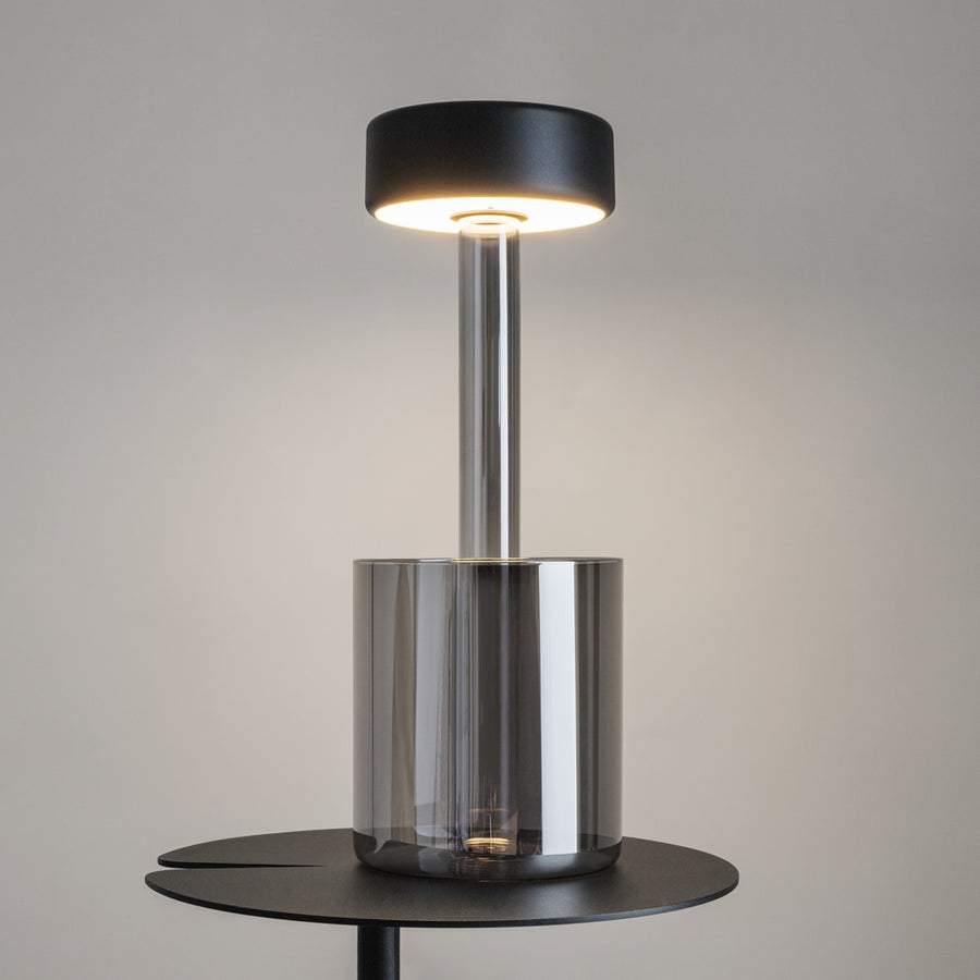 Lampe de chevet grise allumée posée sur table, verre fumé et halo lumineux chaud.