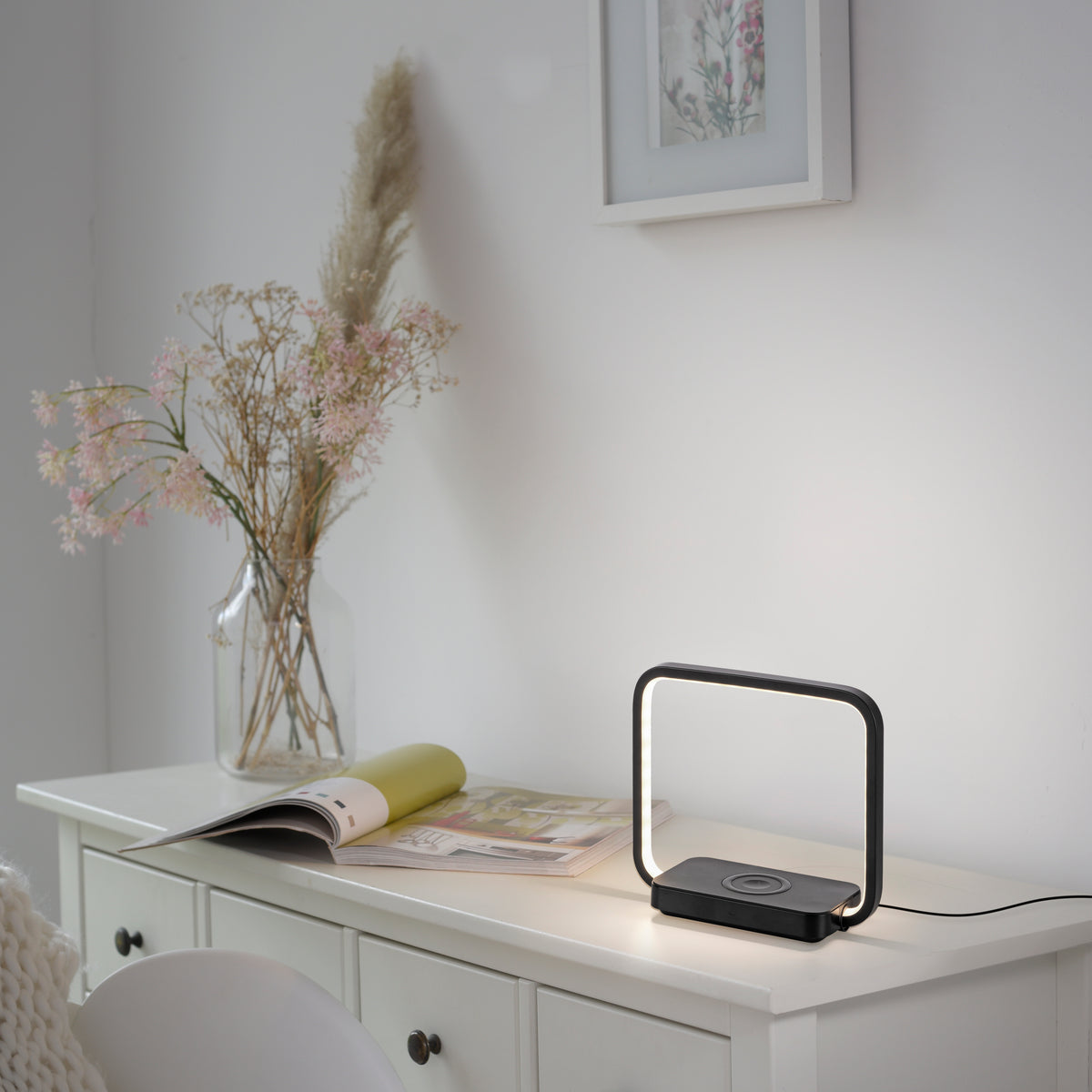 Lampe posée sur une commode blanche, entourée d'un vase de fleurs séchées et d'un magazine.