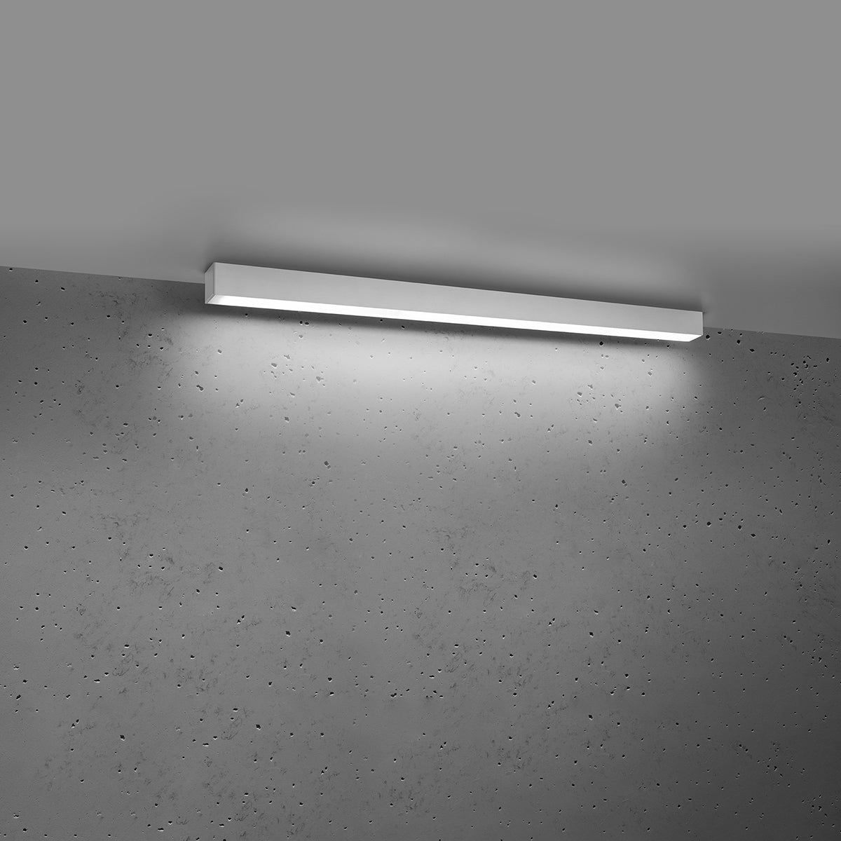 Plafonnier blanc allumé fixé au plafond, éclairant un mur en béton brut.