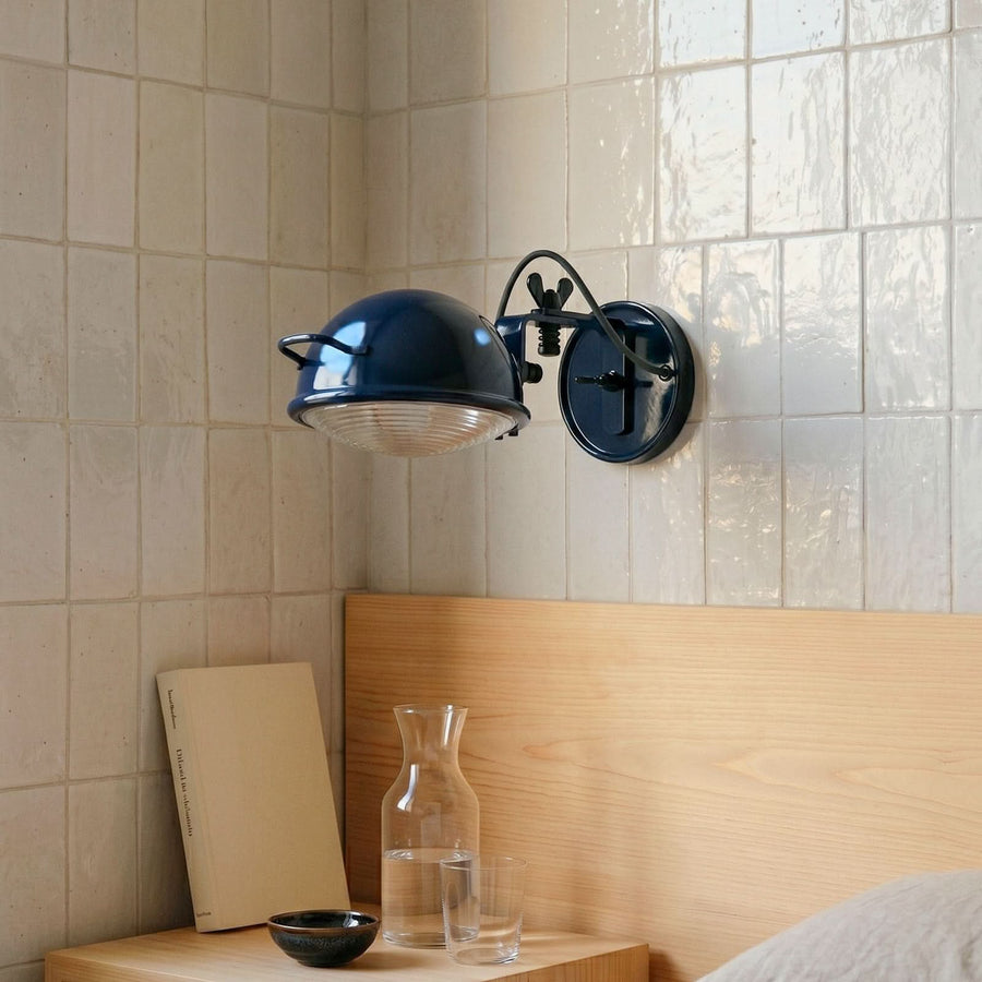 Luminaire mural bleu installé dans une salle de bain carrelée, au-dessus d’un chevet en bois.