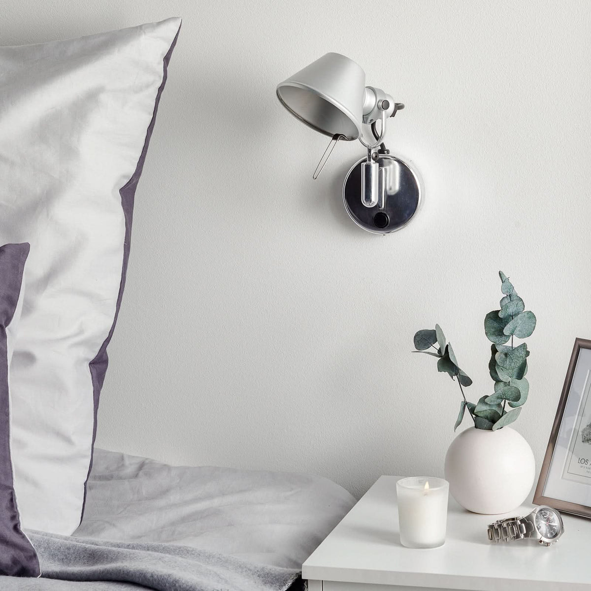 Vue en ambiance d'une applique murale LED fixée près d'un lit gris clair et d'un chevet.