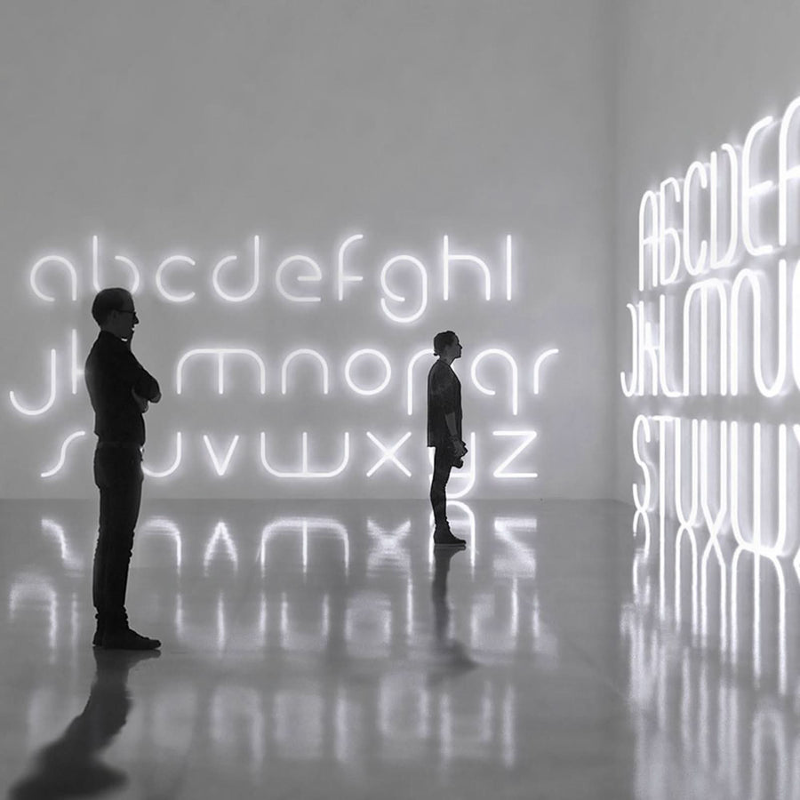 Deux silhouettes contemplent un alphabet lumineux blanc installé sur les murs d'une galerie.