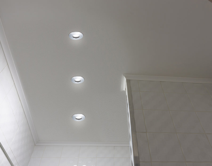 Trois spots encastrés allumés alignés au plafond blanc d'une salle de bain carrelée