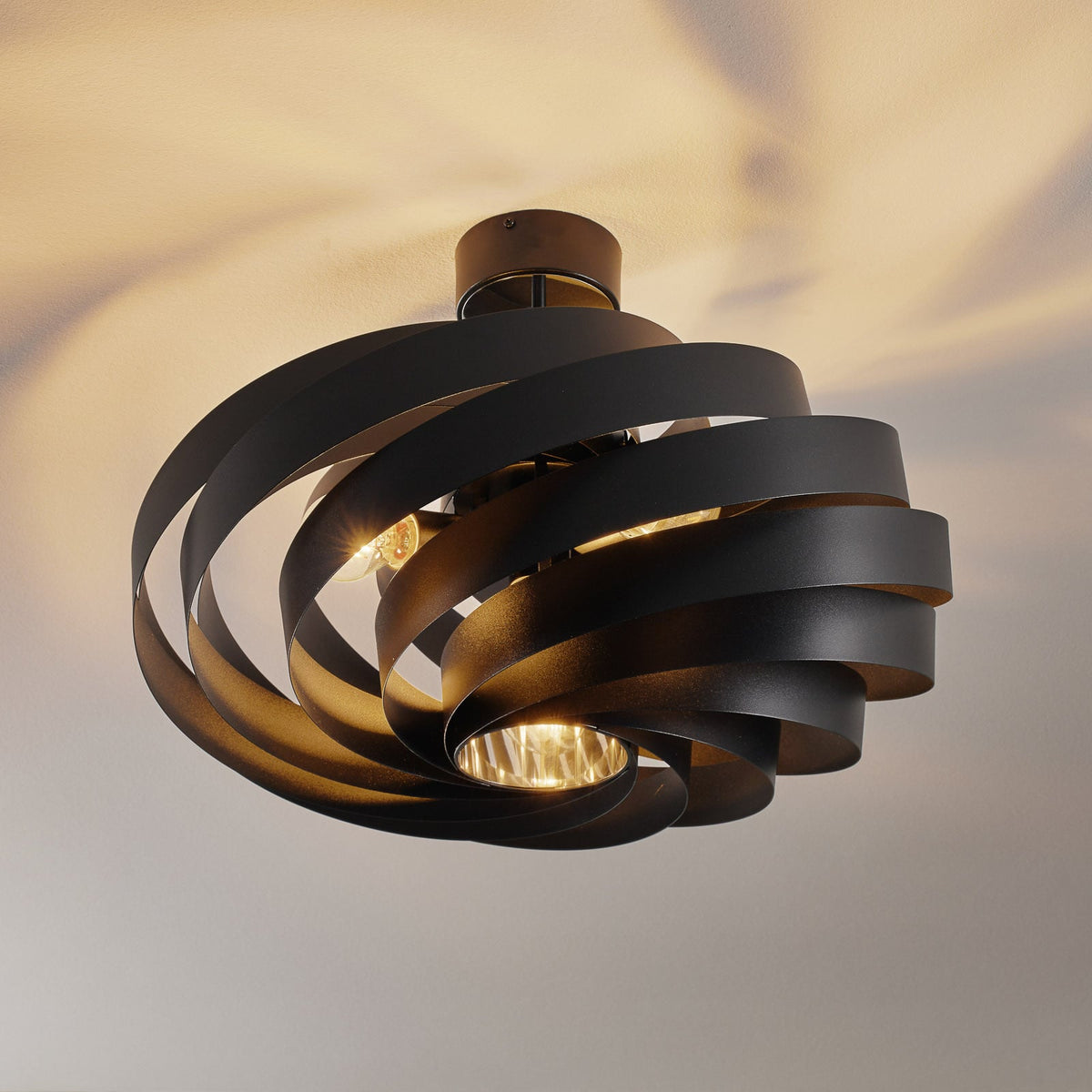 Plafonnier noir allumé, fixé au plafond, diffusant une lumière chaude et des ombres portées.