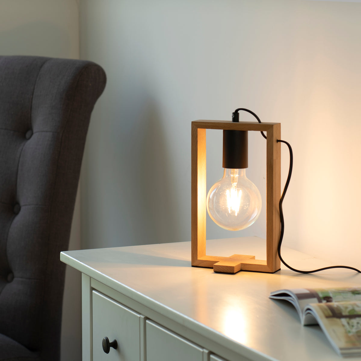 Lampe allumée posée sur une commode blanche près d'un fauteuil gris, ambiance chaleureuse.
