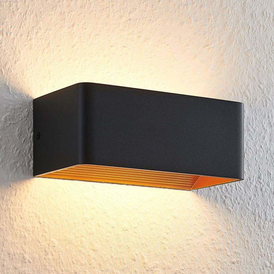 Gros plan de l'applique allumée, grille dorée sous le boîtier noir et lumière chaude.
