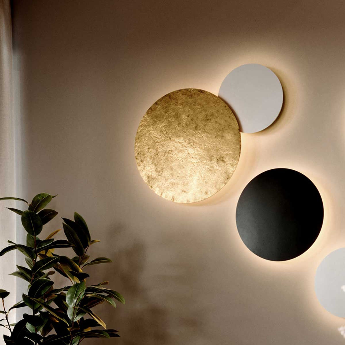 Détail de trois disques muraux aux finitions dorée, noire et blanche sur mur beige.