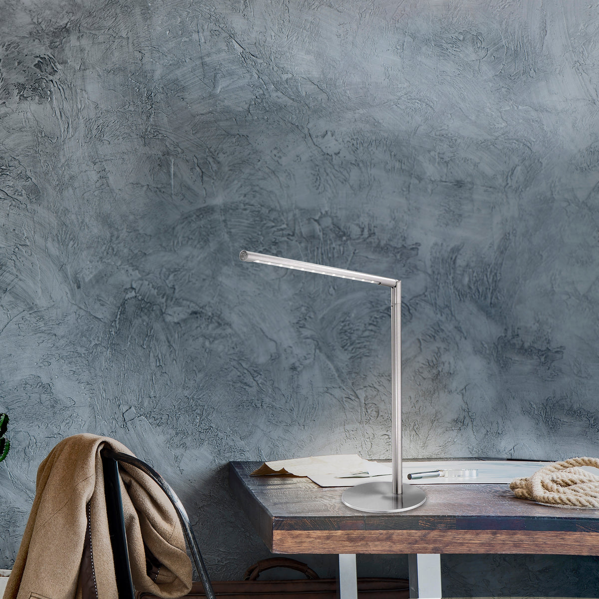 Mise en ambiance sur un bureau en bois, lampe argentée allumée devant un mur béton gris.