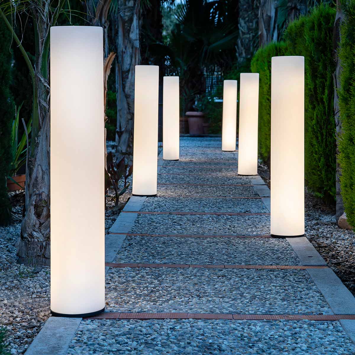 Alignement de six lampadaires cylindriques lumineux bordant une allée de gravier dans un jardin