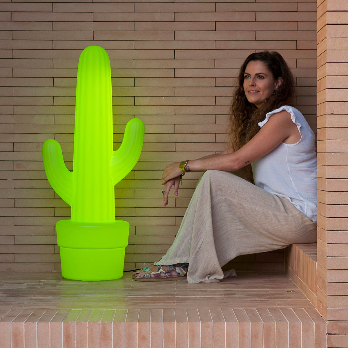 Femme assise près du lampadaire cactus vert, sur marches carrelées, lumière éteinte.