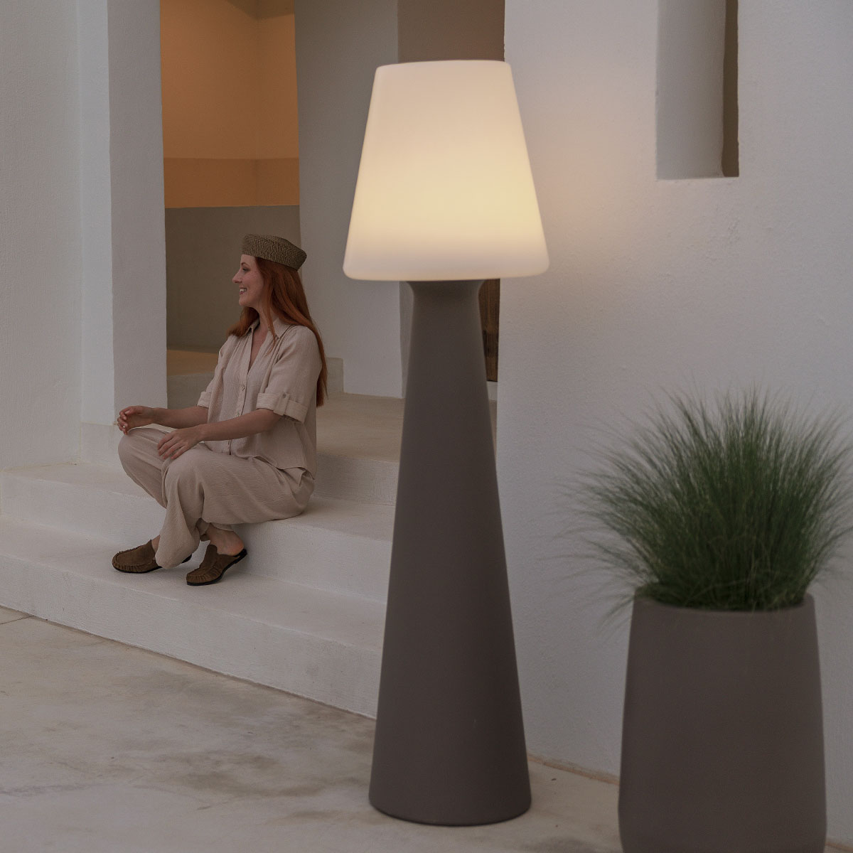 Mise en ambiance : femme assise sur marches blanches près du lampadaire allumé et d'une plante.