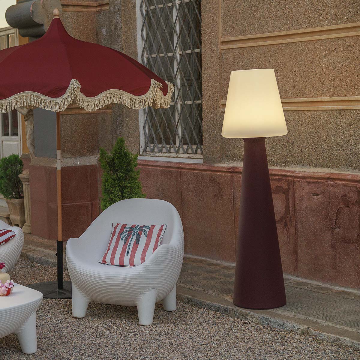 Lampadaire marron foncé installé sur une terrasse en gravier près d'un parasol rouge.