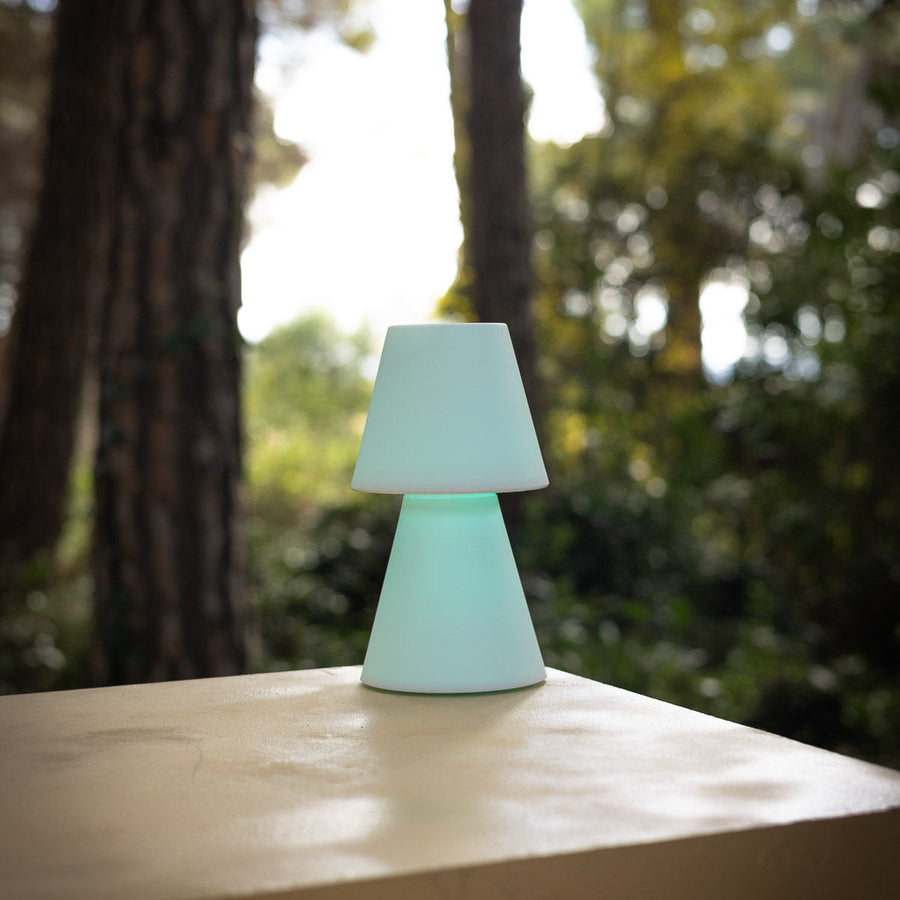 Lampe à poser blanche mate posée sur une table en extérieur, arbres flous en arrière-plan.