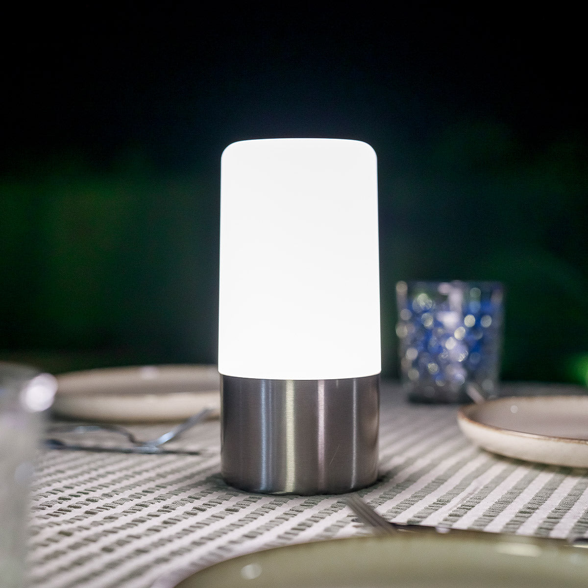 Vue rapprochée du luminaire posé sur nappe rayée, base acier brossé et cylindre blanc.