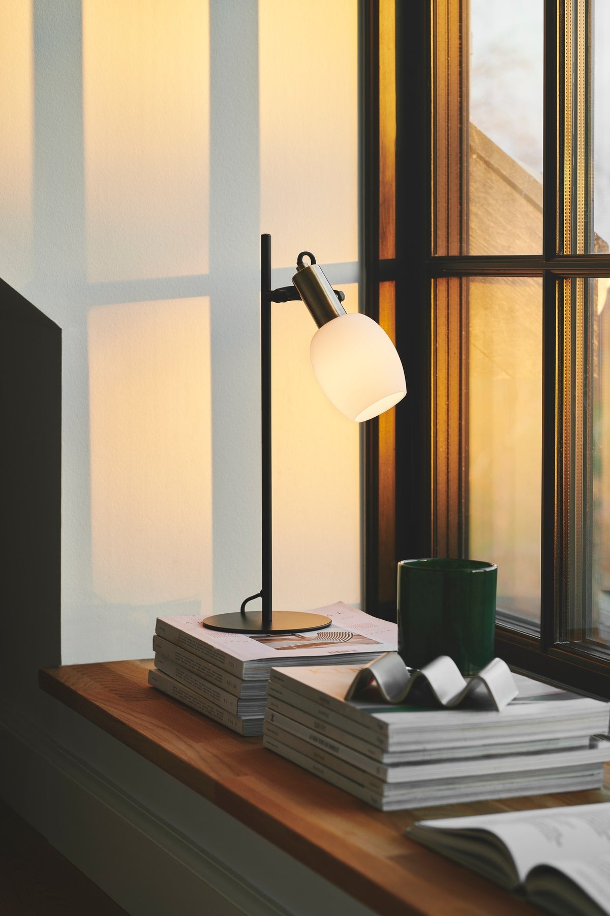 Mise en ambiance : lampe allumée près de piles de livres sur un rebord ensoleillé.
