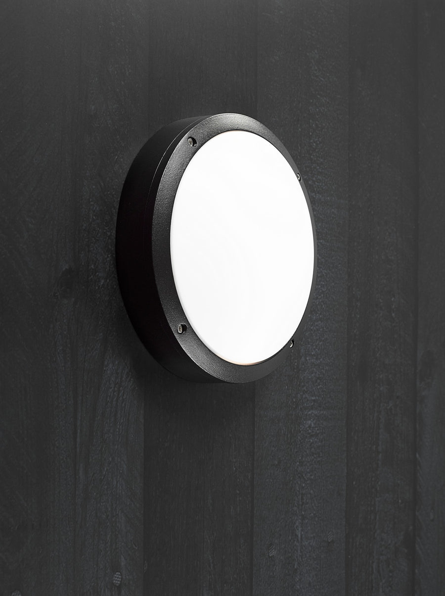Luminaire rond noir installé sur une paroi en bois vertical, éclairant vers l'extérieur.