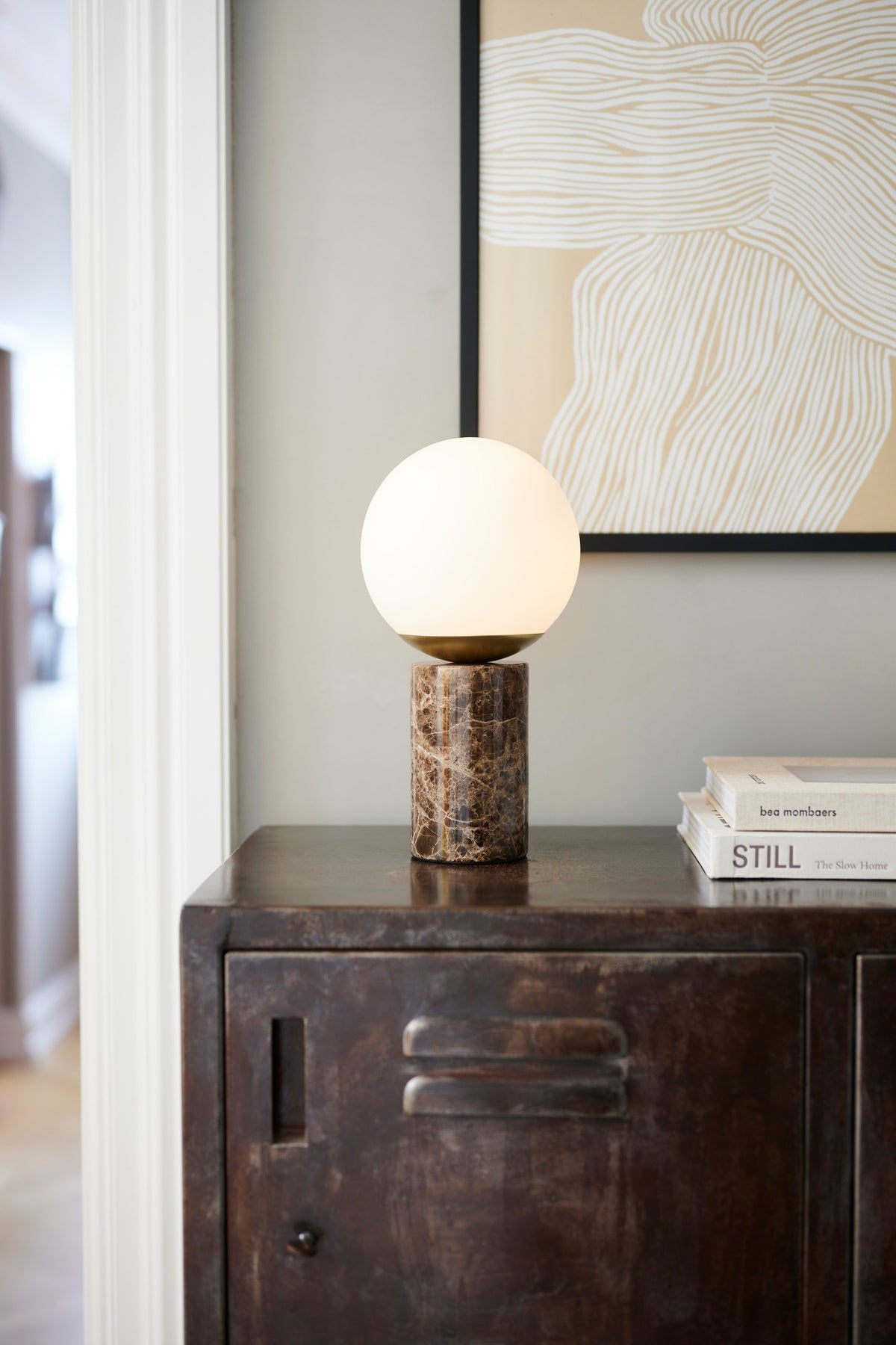 Mise en ambiance : lampe allumée posée sur un meuble sombre devant un cadre.