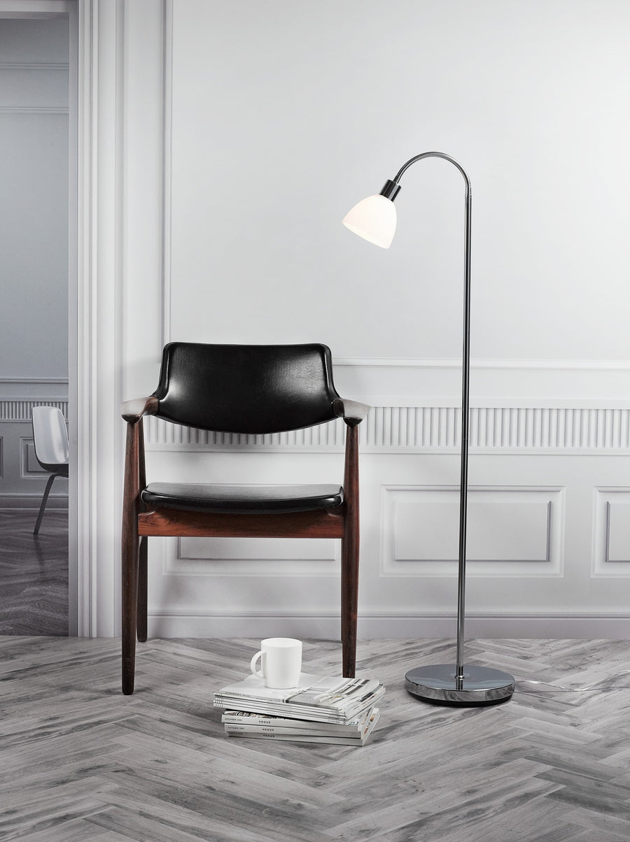 Lampadaire chromé allumé près d'un fauteuil noir dans un intérieur gris.
