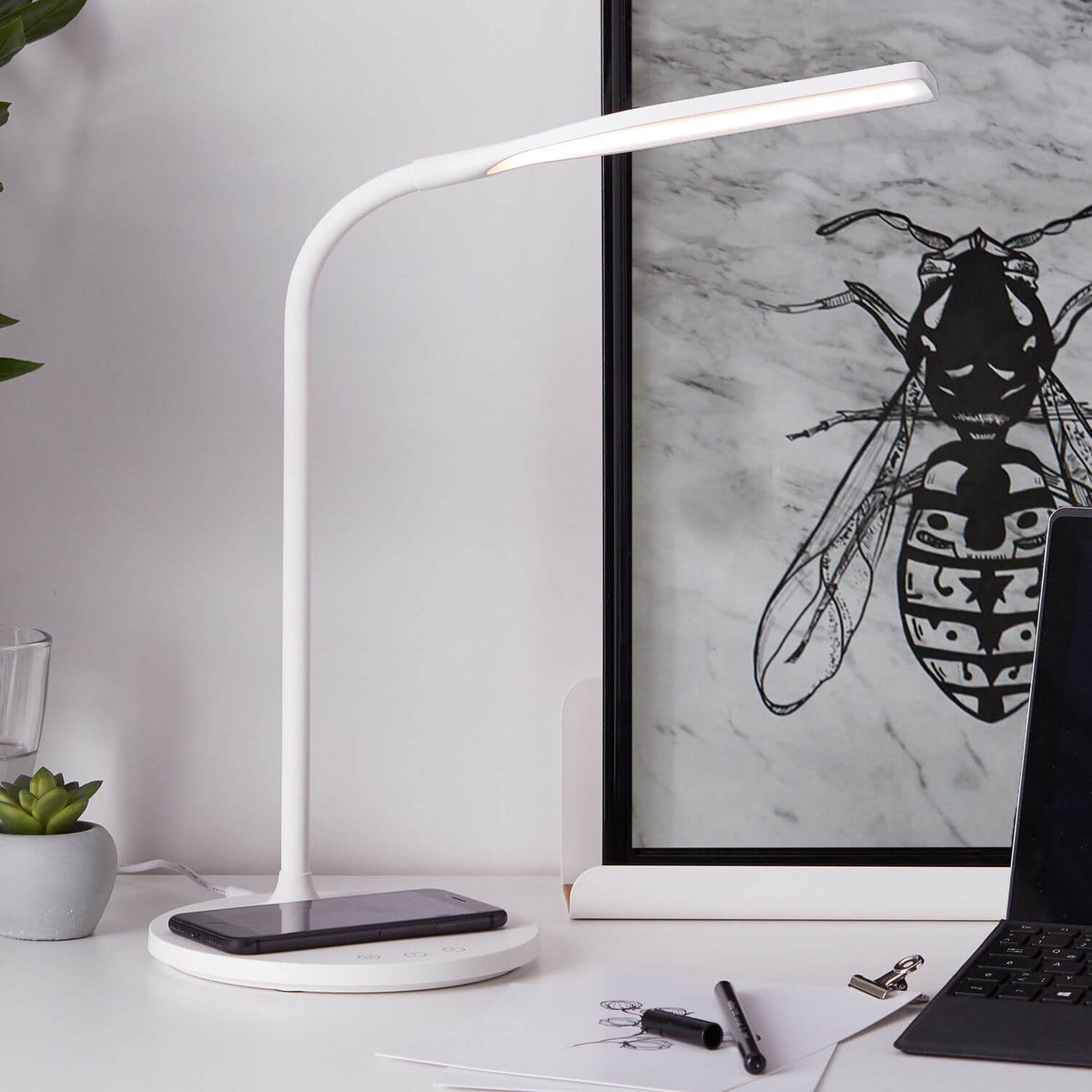Lampe posée sur un bureau, smartphone en recharge sans fil devant une affiche encadrée