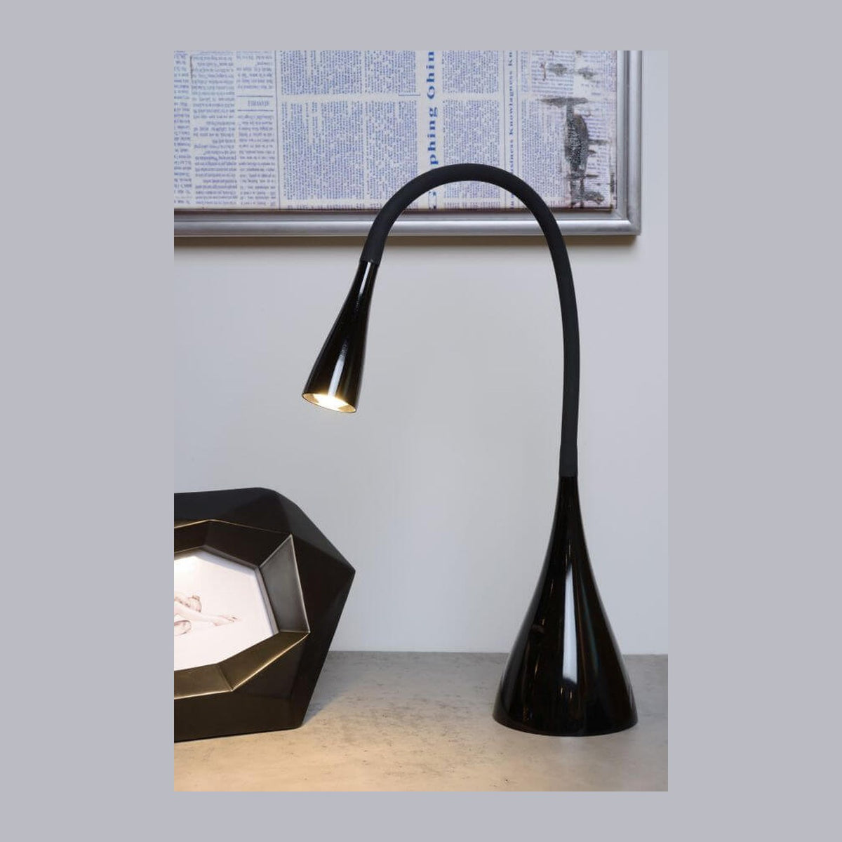 Lampe de bureau noire allumée posée sur une table, cadre journal en arrière-plan