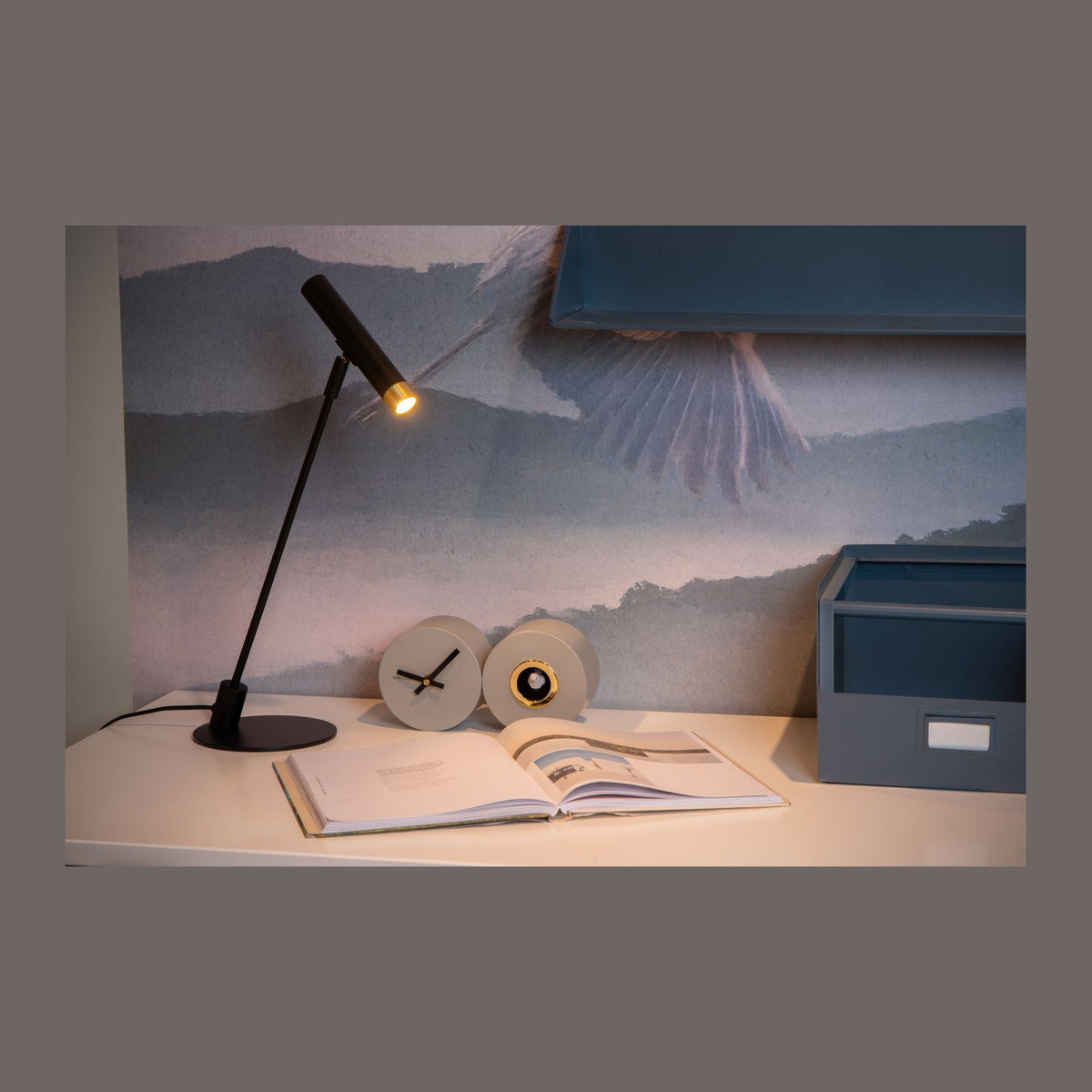 Lampe noire allumée posée sur un bureau blanc, éclairant un livre ouvert près d'un réveil
