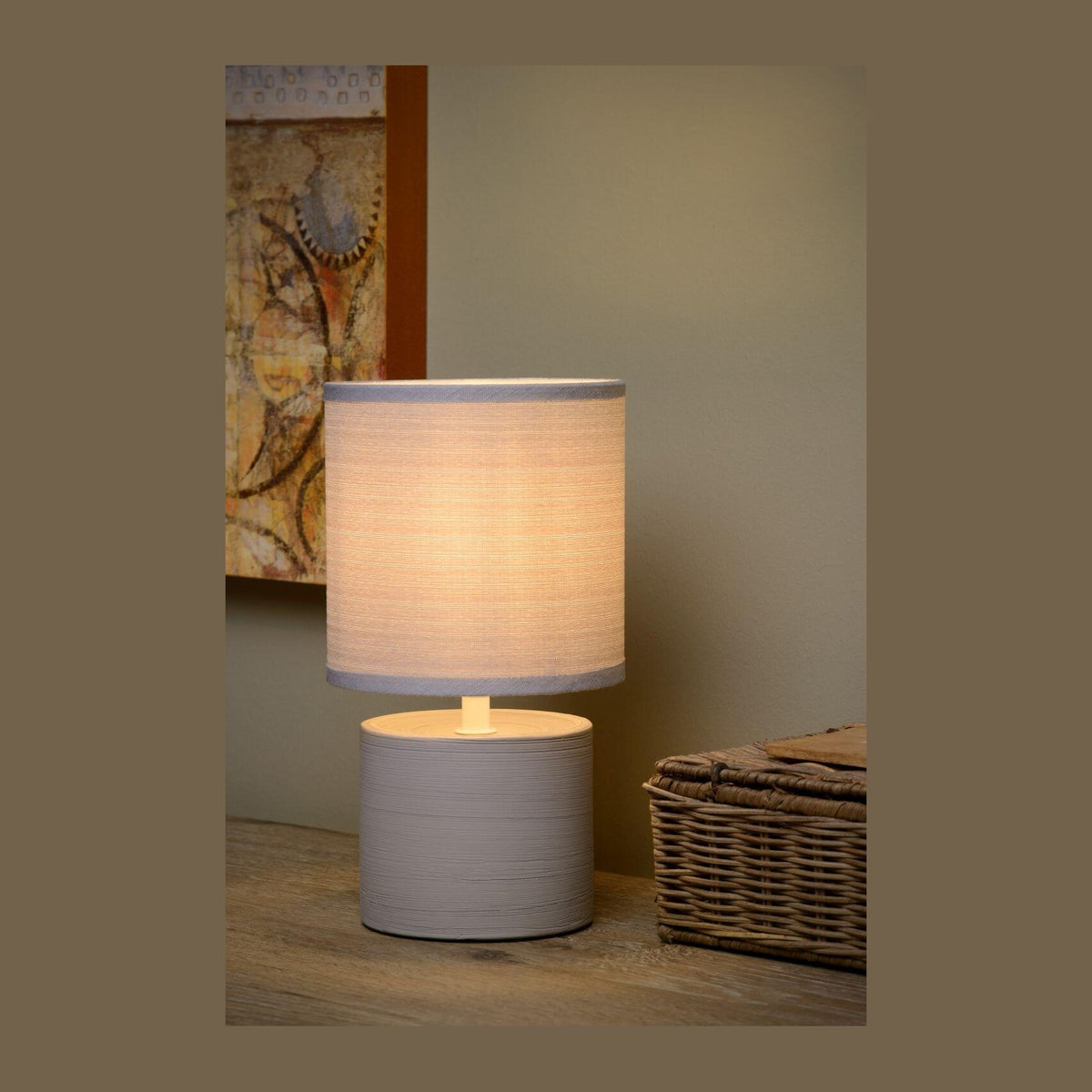 Lampe à poser grise allumée sur une table en bois, panier tressé et tableau en arrière-plan.