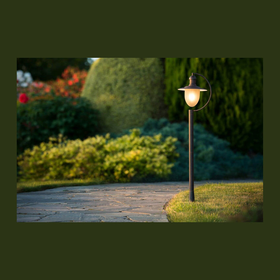 Lanterne sur mât rouille allumée, vue de face dans un jardin paysager.