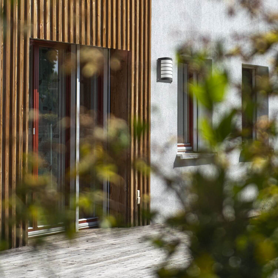 Applique murale anthracite installée sur une façade moderne, avec fenêtres et feuillage flou au premier plan.