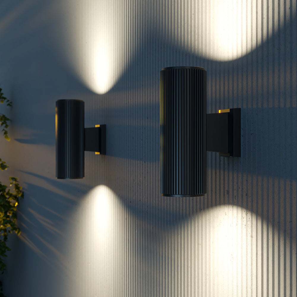 Deux appliques noires allumées sur un mur strié, ombres portées et feuillage