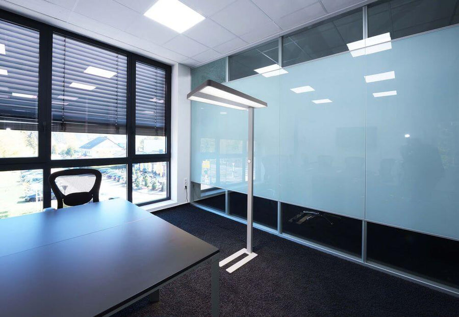 Lampadaire installé dans un bureau, près d'une table et de grandes fenêtres à stores.