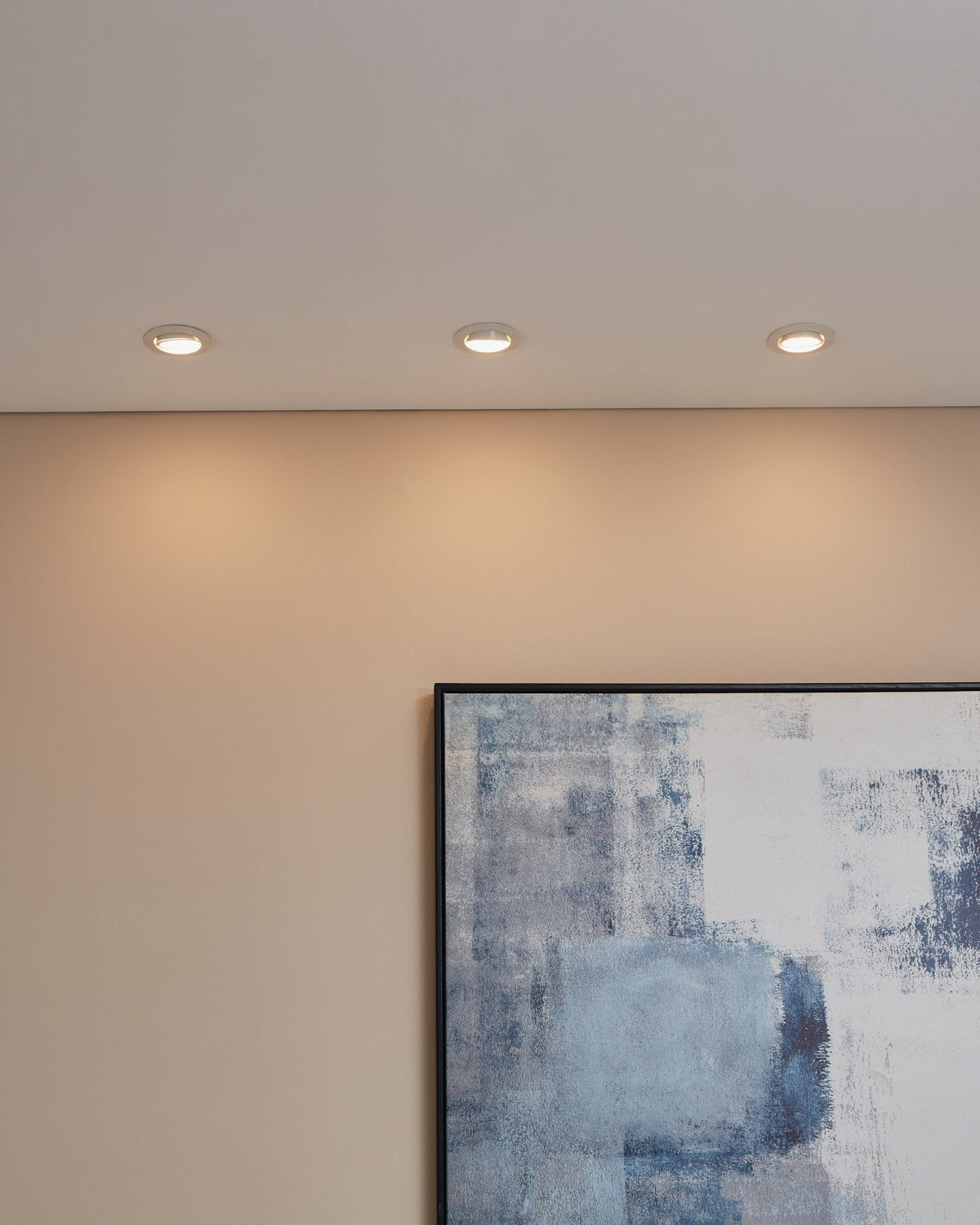 Trois spots encastrés au plafond éclairant un mur beige et un tableau